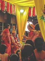 A wedding ceremony with several people dressed in traditional attire. The setting includes vibrant colors with yellow drapes and red and white flower decorations. A man is seated on a throne-like chair wearing ceremonial clothing and a turban, while a woman in a red saree stands nearby. The ambiance is festive, with other people surrounding the scene.