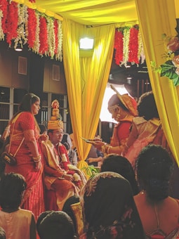 A wedding ceremony with several people dressed in traditional attire. The setting includes vibrant colors with yellow drapes and red and white flower decorations. A man is seated on a throne-like chair wearing ceremonial clothing and a turban, while a woman in a red saree stands nearby. The ambiance is festive, with other people surrounding the scene.