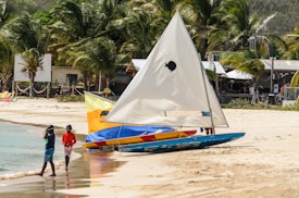 A sandy beach scene features two children walking near the shoreline beside a colorful sailboat, which rests on the sand. Behind them, lush palm trees line the beach, and a few buildings are visible in the background, giving a sense of a lively yet relaxed atmosphere.