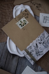 A collection of intricately designed vintage envelopes and papers arranged on a wooden surface. The envelopes feature floral and botanical motifs with handwritten text, and some have decorative stamps. A white ceramic dish is partially visible beneath the documents.