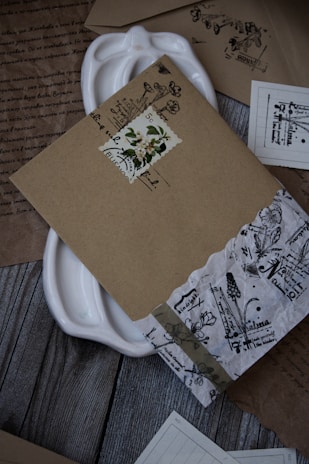 A collection of intricately designed vintage envelopes and papers arranged on a wooden surface. The envelopes feature floral and botanical motifs with handwritten text, and some have decorative stamps. A white ceramic dish is partially visible beneath the documents.