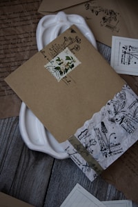A collection of intricately designed vintage envelopes and papers arranged on a wooden surface. The envelopes feature floral and botanical motifs with handwritten text, and some have decorative stamps. A white ceramic dish is partially visible beneath the documents.