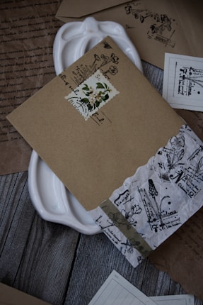 A collection of intricately designed vintage envelopes and papers arranged on a wooden surface. The envelopes feature floral and botanical motifs with handwritten text, and some have decorative stamps. A white ceramic dish is partially visible beneath the documents.
