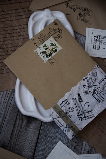 A collection of intricately designed vintage envelopes and papers arranged on a wooden surface. The envelopes feature floral and botanical motifs with handwritten text, and some have decorative stamps. A white ceramic dish is partially visible beneath the documents.