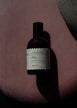 A bottle of perfume labeled 'Floral/Frutal' placed on a soft, textured pink surface with an abstract grey shape overlapping on the right-hand side. The lighting creates a shadow of the bottle, enhancing the contrast and giving a dramatic effect.