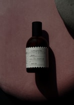 A bottle of perfume labeled 'Floral/Frutal' placed on a soft, textured pink surface with an abstract grey shape overlapping on the right-hand side. The lighting creates a shadow of the bottle, enhancing the contrast and giving a dramatic effect.