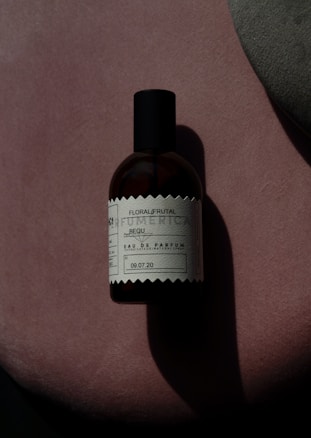 A bottle of perfume labeled 'Floral/Frutal' placed on a soft, textured pink surface with an abstract grey shape overlapping on the right-hand side. The lighting creates a shadow of the bottle, enhancing the contrast and giving a dramatic effect.