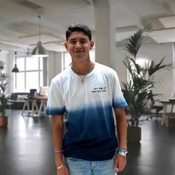 A young man standing in a modern, well-lit office space, wearing a white and blue gradient t-shirt with the text 'GET RID OF THE ROUTINE'. The background features office furniture and potted plants, creating a professional yet relaxed atmosphere.