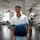 A young man standing in a modern, well-lit office space, wearing a white and blue gradient t-shirt with the text 'GET RID OF THE ROUTINE'. The background features office furniture and potted plants, creating a professional yet relaxed atmosphere.