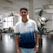 A young man standing in a modern, well-lit office space, wearing a white and blue gradient t-shirt with the text 'GET RID OF THE ROUTINE'. The background features office furniture and potted plants, creating a professional yet relaxed atmosphere.
