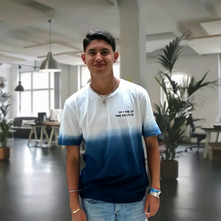 A young man standing in a modern, well-lit office space, wearing a white and blue gradient t-shirt with the text 'GET RID OF THE ROUTINE'. The background features office furniture and potted plants, creating a professional yet relaxed atmosphere.