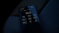 A smartphone screen displaying detailed battery-related statistics in German, such as voltage, temperature, and range. The phone is placed on a dark textured fabric surface, possibly a car seat.