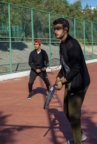 Two individuals are on a tennis court, each holding a tennis racket. They appear to be in a ready stance, poised to play. The court is surrounded by a green metal fence, and natural greenery can be seen in the background.