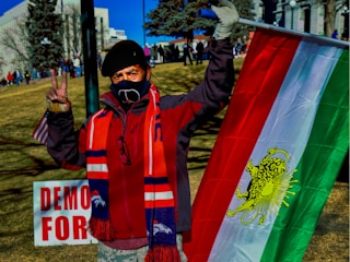 A person wearing a mask and a beret holds a flag with red, white, and green stripes with a lion symbol. The individual gestures a peace sign with one hand and stands in a park setting with several other people in the background. A sign partially reads 'DEMO' and 'FOR' in red letters.