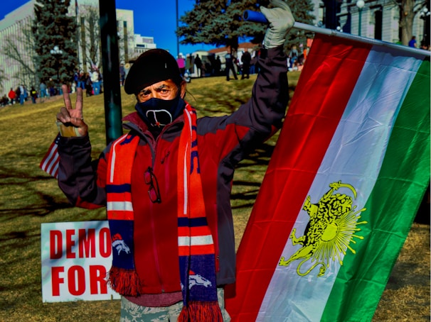 A person wearing a mask and a beret holds a flag with red, white, and green stripes with a lion symbol. The individual gestures a peace sign with one hand and stands in a park setting with several other people in the background. A sign partially reads 'DEMO' and 'FOR' in red letters.