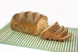 A loaf of brown bread with slices cut and laid out beside it on a green and white striped mat. The bread appears to have a crusty texture with notable cuts on the top for design.