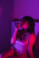 A person is sitting and holding a cigarette with smoke swirling above. The lighting is low and dramatic, featuring deep shades of pink and purple, casting vivid shadows on the figure.