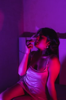 A person is sitting and holding a cigarette with smoke swirling above. The lighting is low and dramatic, featuring deep shades of pink and purple, casting vivid shadows on the figure.