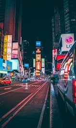 A vibrant, bustling city street at night with bright digital billboards and advertising signs illuminating the scene. Vehicles are visible on the road, including a parked van on the right. Skyscrapers and buildings line the avenue, showcasing colorful light displays.