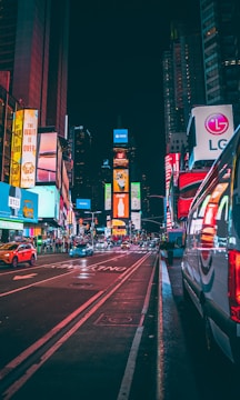 A vibrant, bustling city street at night with bright digital billboards and advertising signs illuminating the scene. Vehicles are visible on the road, including a parked van on the right. Skyscrapers and buildings line the avenue, showcasing colorful light displays.