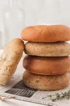 A stack of three mixed-grain bagels is arranged on top of a newspaper. One bagel is leaning against the stack, showcasing a speckled texture. The background is soft and blurred, with hints of a glass bottle and a sprig of herbs adding to the aesthetic.