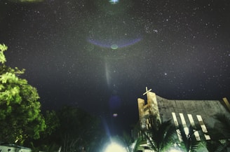 A starry night sky filled with twinkling stars above a church with a visible cross at the top. The silhouette of trees is visible at the bottom, with a mix of dark and illuminated leaves. The sky features some lens flares and possibly planets or celestial objects, adding a mystic quality to the scene.