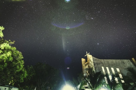 A starry night sky filled with twinkling stars above a church with a visible cross at the top. The silhouette of trees is visible at the bottom, with a mix of dark and illuminated leaves. The sky features some lens flares and possibly planets or celestial objects, adding a mystic quality to the scene.