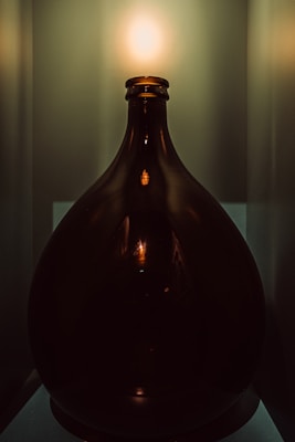 A large, dark glass bottle is positioned on a surface, accentuated by a soft, warm glow emanating from above. The light creates a reflection on the bottle's glossy surface, highlighting its curved silhouette. The background is muted with a gradient of green hues, enhancing the focus on the bottle.