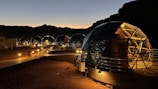 A series of geodesic dome structures are illuminated against a dusky sky, positioned along a wooden pathway in a desert setting. The domes have transparent panels revealing a warm interior light, suggesting a cozy and modern accommodation. The surrounding landscape is marked by silhouetted mountain ridges, creating a tranquil and remote atmosphere.