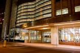 A well-lit, modern building entrance with large glass windows. The structure's facade showcases a combination of metal and concrete with illuminated signage. Several American flags are positioned near the entrance, and the interior appears welcoming with visible seating arrangements.