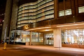 A well-lit, modern building entrance with large glass windows. The structure's facade showcases a combination of metal and concrete with illuminated signage. Several American flags are positioned near the entrance, and the interior appears welcoming with visible seating arrangements.