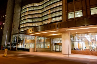 A well-lit, modern building entrance with large glass windows. The structure's facade showcases a combination of metal and concrete with illuminated signage. Several American flags are positioned near the entrance, and the interior appears welcoming with visible seating arrangements.