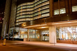 A well-lit, modern building entrance with large glass windows. The structure's facade showcases a combination of metal and concrete with illuminated signage. Several American flags are positioned near the entrance, and the interior appears welcoming with visible seating arrangements.