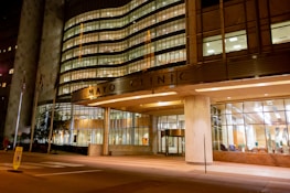 A well-lit, modern building entrance with large glass windows. The structure's facade showcases a combination of metal and concrete with illuminated signage. Several American flags are positioned near the entrance, and the interior appears welcoming with visible seating arrangements.