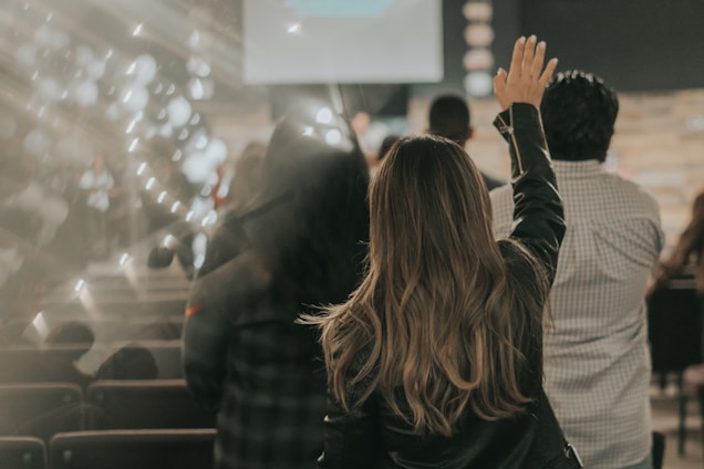 A gathering of people in what appears to be an indoor event or seminar, with blurred lights providing a dreamy effect. A person prominently stands with a raised hand in the foreground, amidst other attendees seated in rows.
