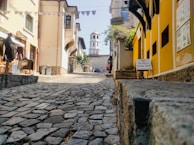 A narrow cobblestone street is flanked by colorful buildings, culminating in a domed, historic structure at the end of the path. Signs indicate a bed and breakfast, adding to the quaint, inviting atmosphere of the area. Small details like hanging banners and potted plants contribute to a charming, rustic vibe.