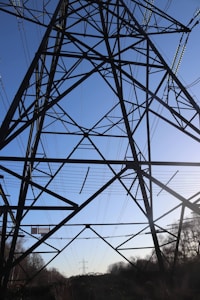 A large metal electricity pylon with an intricate lattice structure stands prominently against a clear blue sky. Numerous cables stretch out from the tower, indicating the transmission of electricity across the landscape. The ground beneath is shadowed, with silhouettes of trees and vegetation visible.