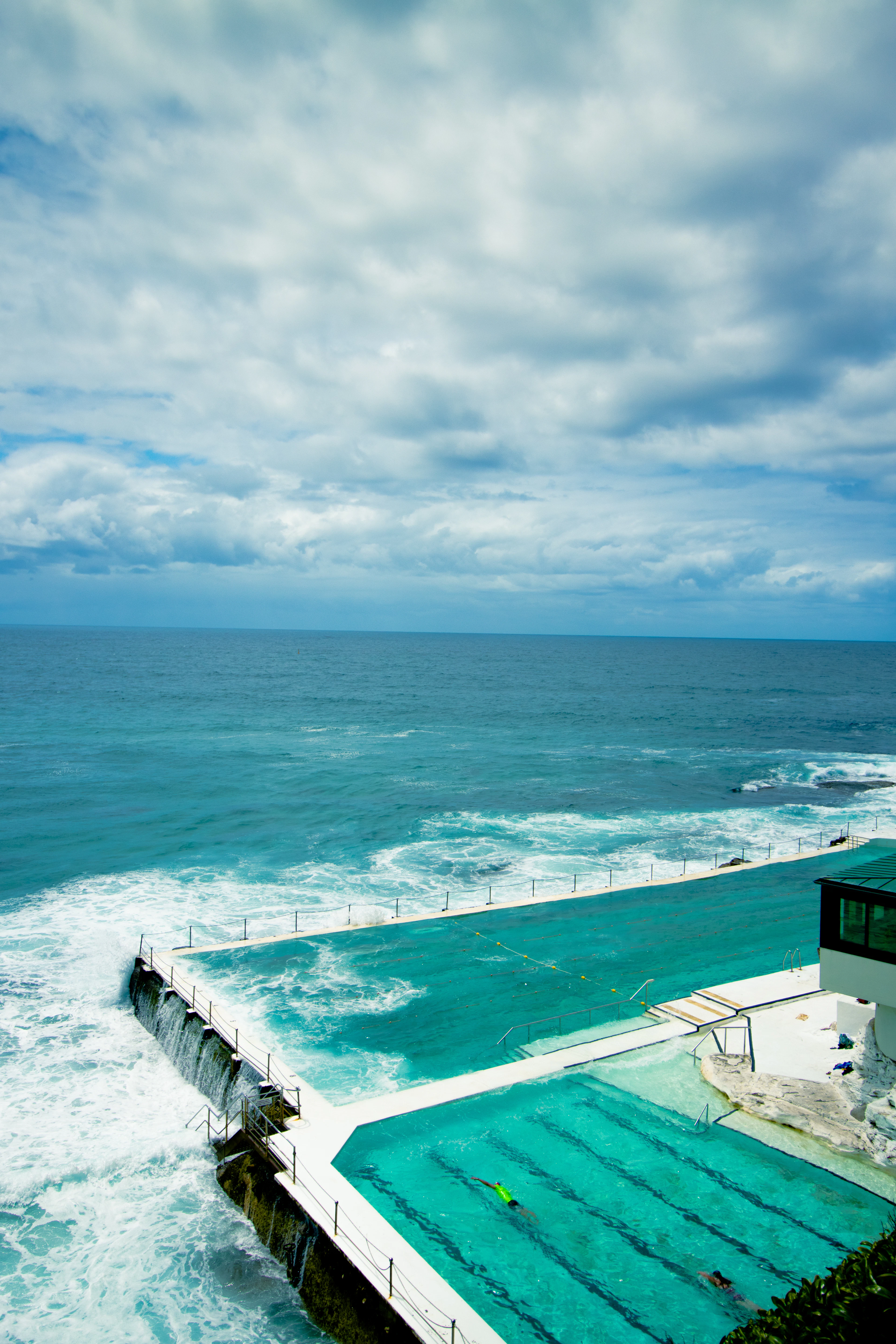 The Bondi Icebergs ocean pool at the edge of the Pacific in Sydney, surf breaking over its sea wall under a cloudy sky