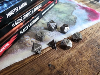 A collection of tabletop role-playing game manuals is stacked neatly on the left side. Next to them, a set of metallic polyhedral dice is arranged on a wooden table, with a colorful map-like cloth underneath.