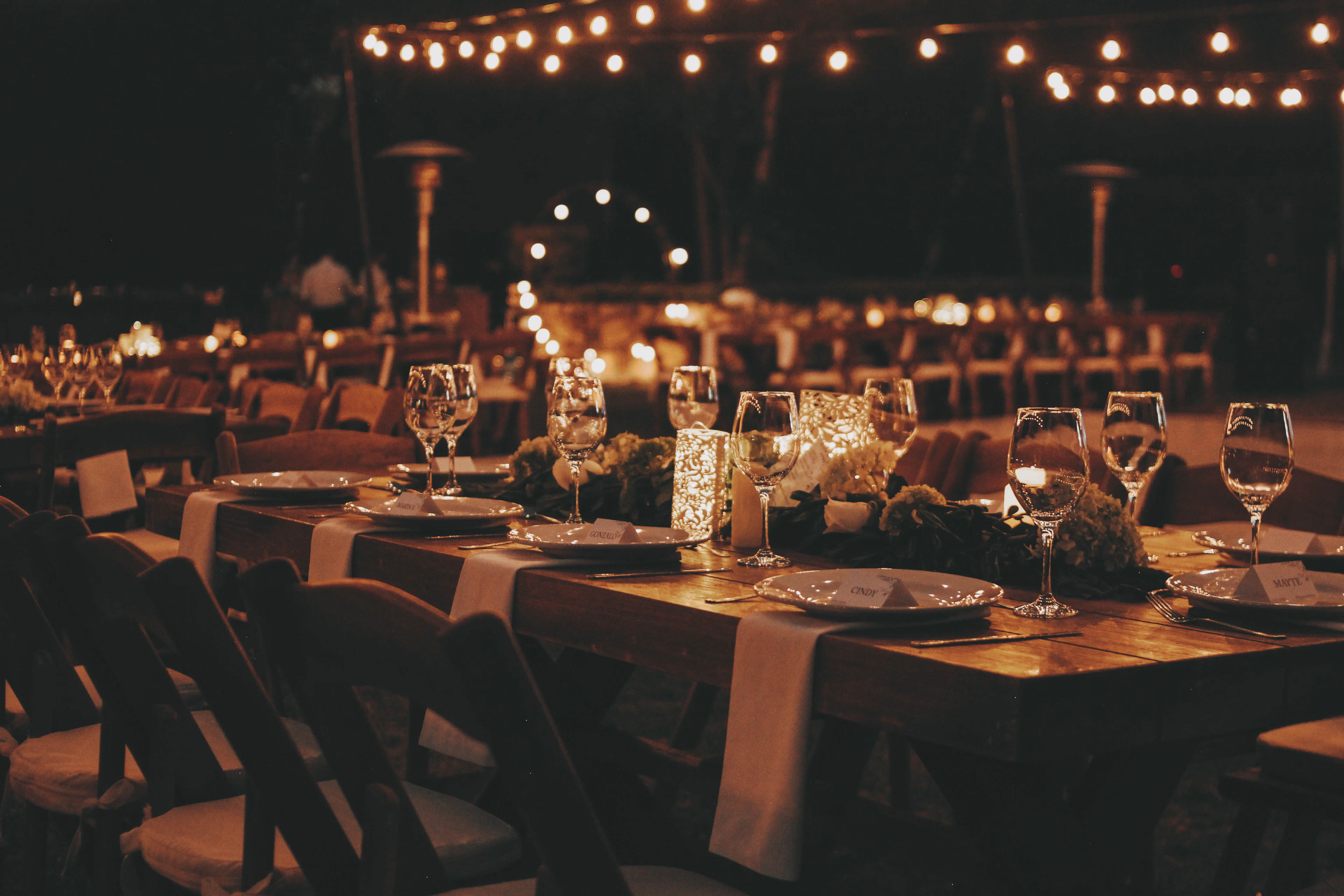 De longues tables en bois sous des guirlandes lumineuses, le soir