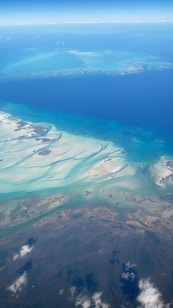 Aerial view of a coastal landscape featuring vibrant blues and turquoise waters, with swirling sandbanks and small islands. The horizon shows the vast expanse of the ocean meeting the sky, with thin, wispy clouds visible in the upper part of the image.