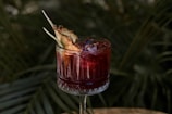A drink in a glass with an intricate design, filled with a red liquid and ice cubes. The rim of the glass is garnished with a slice of pineapple. The background consists of green, leafy plants, giving an outdoor and tropical vibe.