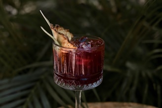 A drink in a glass with an intricate design, filled with a red liquid and ice cubes. The rim of the glass is garnished with a slice of pineapple. The background consists of green, leafy plants, giving an outdoor and tropical vibe.