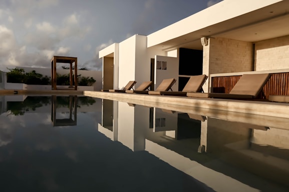 An outdoor pool beside a modern, minimalist building with clean, geometric lines. The pool reflects the sky and nearby structures, creating a mirror-like effect. Several wooden deck chairs with cushions are arranged in a row beside the pool. Foliage can be seen in the background under a partly cloudy sky, adding a touch of nature to the modern setting.