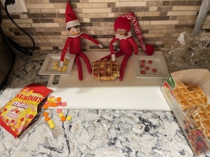 A festive Elf on the Shelf setup on a kitchen counter surrounded by family snacks and crafts