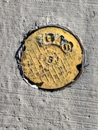 A round, weathered yellow metal marker embedded in rough concrete. It has the word 'GAS' and a number '5' prominently engraved, with some patches where the paint has eroded.