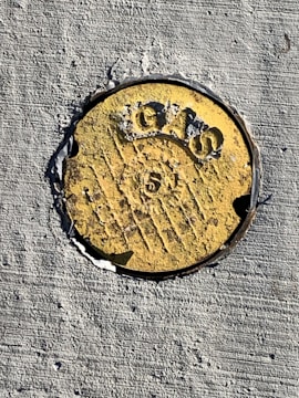 A round, weathered yellow metal marker embedded in rough concrete. It has the word 'GAS' and a number '5' prominently engraved, with some patches where the paint has eroded.
