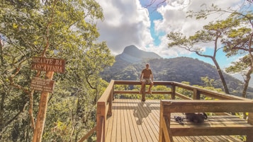 A wooden viewing platform overlooking a lush, forested mountain landscape with a prominent peak in the background. A person sits on the railings of the platform, and there's a wooden sign to the left that features the words 'Mirante da Cascatinha.' The sky is partly cloudy, allowing sunlight to filter through and illuminate the scene.
