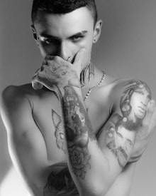 A person with a short haircut and piercings is covering their mouth with their hand. Numerous tattoos are visible on the arms and neck, including faces and animals. The lighting is dramatic, casting shadows on the face and body.