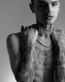 A black and white portrait of a person with tattoos on their arms and chest, including a cat with a third eye on their sternum. They have piercings and are holding the collar of a shirt, revealing intricate neck tattoos. The image has a shadowy and dramatic lighting.